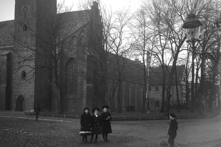 Svartvitt foto från tidigt 1900-tal. Tre flickor och en pojke står mittemot varandra på kullerstensgata framför klostret och höga träd. 