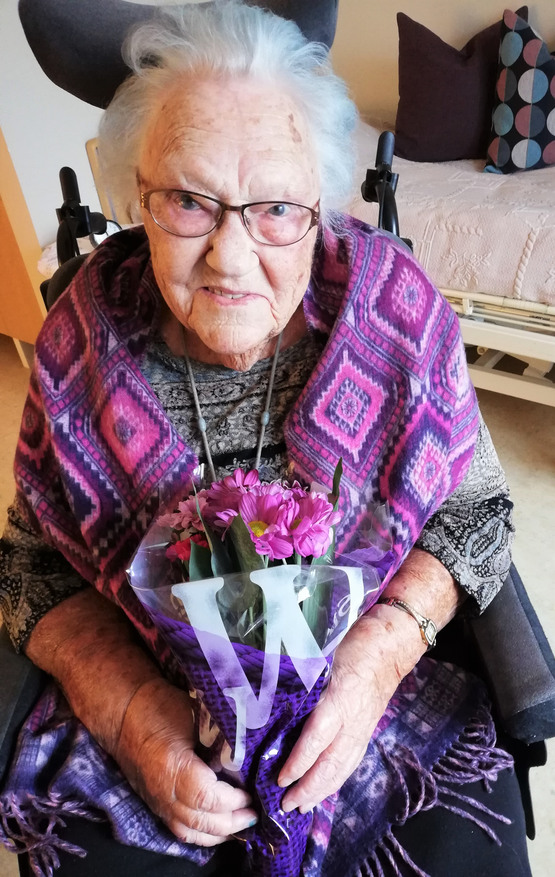 Äldre kvinna i lilamönstras sjal håller en bukett lila blommor.